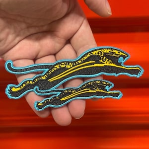 May include: Two embroidered patches featuring a leaping jaguar design. The jaguars are black with yellow stripes and a light blue outline. One patch is larger than the other.