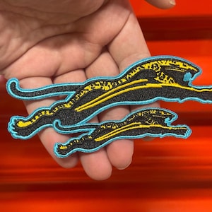 May include: Two embroidered patches featuring a leaping jaguar design. The patches have a black background with yellow and blue accents. The jaguar's body is black with yellow spots, outlined in blue. The patches are in different sizes.