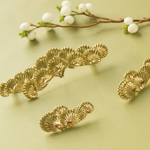 May include: Three gold-toned metal drawer pulls shaped like stylized fan leaves. The pulls are arranged in a row, with the largest pull in the center and the two smaller pulls on either side.