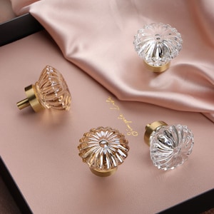 Op de afbeelding: Vier glazen kastknoppen met een gouden metalen basis. De knoppen hebben een heldere en amberkleurige glazen afwerking met een getextureerd, bloemvormig ontwerp.