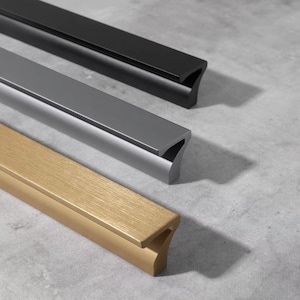 May include: Three cabinet pulls in different finishes: black, grey, and gold. Each pull has a rectangular shape with a curved edge on one side.