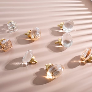 Op de afbeelding: Assortiment van heldere en amberkleurige glazen kastknoppen met gouden metalen bases. De knoppen zijn in verschillende vormen, waaronder rond, ovaal en vierkant.