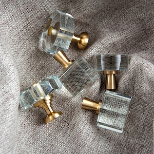 Peut inclure: Plusieurs boutons de placard en cristal transparent avec des bases dorées. Les boutons présentent des surfaces facettées et des formes géométriques variées. Ils sont disposés sur un tissu texturé de ton neutre, pour un effet luxueux.