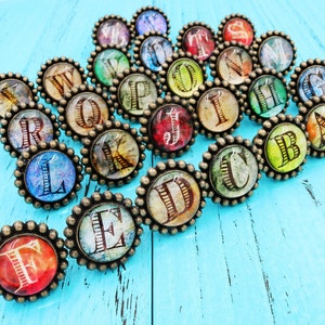 Op de afbeelding: Een verzameling alfabetletterbedels in antieke stijl met een bronzen, parelrand. Elke bedel heeft een andere letter in een vintage lettertype, tegen een kleurrijke, getextureerde achtergrond. De bedels zijn gerangschikt op een helder turkoois oppervlak.