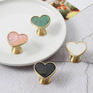 Könnte beinhalten: Vier herzförmige Schrankknöpfe mit Messingbasis und verschiedenen farbigen Einlagen. Die Knöpfe sind grün, rosa, weiß und schwarz.