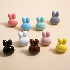Op de afbeelding: Negen keramische trekknopjes in de vorm van een konijn in verschillende kleuren, waaronder roze, blauw, groen, wit, zwart, bruin en geel.