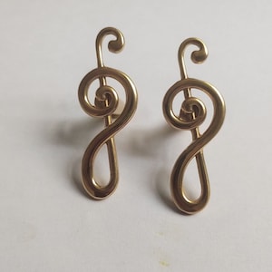 Könnte beinhalten: Zwei goldfarbene Ohrringe in Form eines Violinschlüssels. Die Ohrringe sind in Form eines Violinschlüssels gestaltet und haben eine glatte, glänzende Oberfläche.