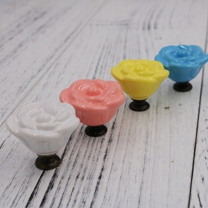 Op de afbeelding: Vier keramische roosvormige ladegrepen in wit, roze, geel en blauw. Elke greep heeft een kleine metalen schroef.