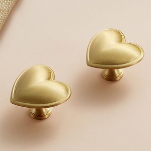 Pode incluir: Dois botões de armário em forma de coração dourados com acabamento escovado.