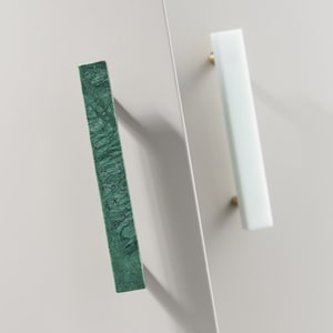 Puede incluir: Dos tiradores de armario, uno verde y otro blanco, con acabado en latón. Los tiradores son rectangulares y tienen una superficie lisa.