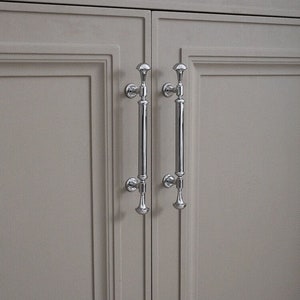 Puede incluir: Dos tiradores de armario plateados con una parte superior redondeada y un mango largo y recto. Los tiradores están montados en una puerta de armario gris claro.