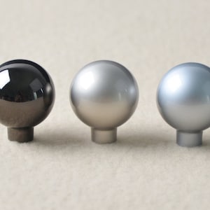 Może przedstawiać: Trzy okrągłe gałki do szafki w różnych wykończeniach: czarny, srebrny i nikiel.