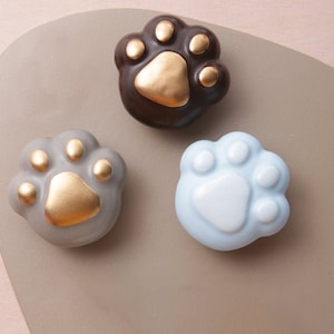 Op de afbeelding: Drie keramische poot afdruk ornamenten in verschillende kleuren: bruin, grijs en lichtblauw. Elke poot afdruk heeft een gouden accent in het midden.