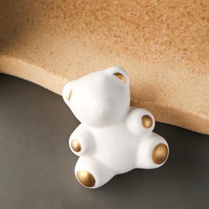 以下が含まれることがあります： 金色のアクセントが付いた白いセラミック製のテディベア人形。クマはシンプルなデザインで、灰色の表面に座っています。