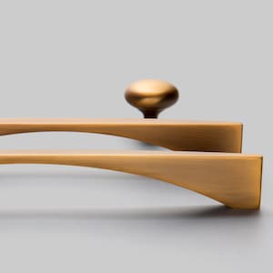 May include: Two brushed gold cabinet pulls with a curved design. The pulls are shown against a grey background.