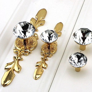 Può includere: Set di quattro pomelli per armadi dorati con maniglie in cristallo trasparente a forma di diamante. I pomelli hanno un design floreale decorativo.
