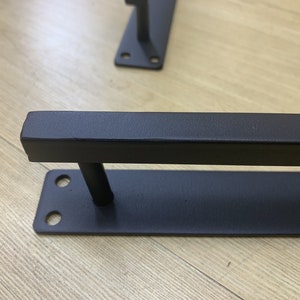 May include: Black metal shelf bracket with two mounting holes. The bracket has a rectangular shape with a raised bar across the top.