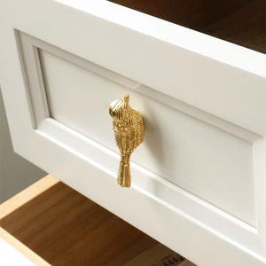 以下が含まれることがあります： 羽根模様が細かく施された金色の鳥の形をした引き出しの取っ手。取っ手は白い引き出しの正面に取り付けられています。