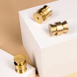 Può includere: Tre pomelli per armadi dorati con un design arrotondato e a strati. I pomelli sono esposti su una superficie bianca.