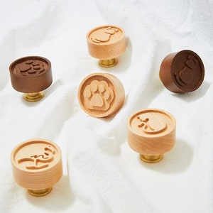 May include: Six wooden cookie stamps with different designs, including a paw print, a cat, and a bird. The stamps are brown and natural wood and have brass accents.