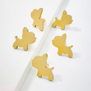 Könnte beinhalten: Fünf goldfarbene Metall-Schrankknöpfe in Form von Cartoon-Hunden. Die Knöpfe befinden sich in verschiedenen Positionen auf einer weißen Oberfläche.