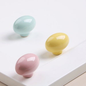 Könnte beinhalten: Drei Keramik-Schrankknöpfe in Pastellfarben: hellblau, gelb und rosa. Die Knöpfe sind oval geformt und haben eine glatte, glänzende Oberfläche.