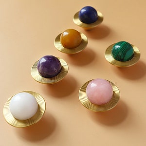 May include: Decorative cabinet knobs with a planetary design. Each knob features a polished, spherical gemstone in various colours, including white, purple, pink, green, yellow, and blue, set on a gold-toned, circular base.