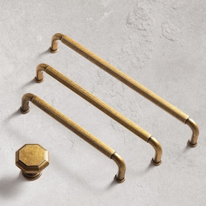 以下が含まれることがあります： 正方形で丸みを帯びたエッジを持つ、アンティークブラスのキャビネットプルが3つあります。プルは、一番上が一番大きく、一番下が一番小さい順に並んでいます。マッチングするアンティークブラスのキャビネットノブも表示されています。