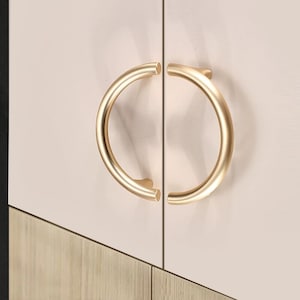 Może przedstawiać: Para złotych uchwytów do szafki o zaokrąglonym, okrągłym kształcie. Uchwyty są zamontowane na jasnoróżowej drzwiach szafki.