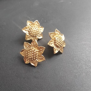 Könnte beinhalten: Drei goldfarbene Ohrringe in Sonnenblumenform. Die Ohrringe sind mit einer strukturierten Mitte verziert und haben einen leicht erhöhten Rand.