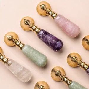 May include: Assortment of six cabinet knobs with gold metal bases and various coloured gemstone-like handles. The handles are in shades of pink, purple, green, and white.