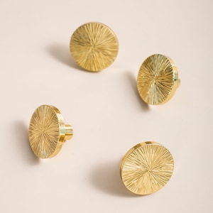 Könnte beinhalten: Vier goldfarbene runde Schrankknöpfe mit einem strukturierten Sonnenstrahl-Design.