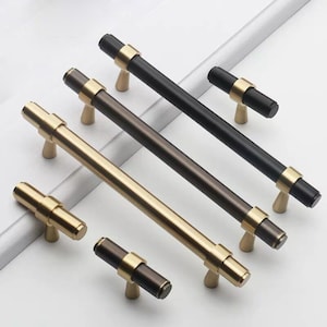 Könnte beinhalten: Fünf Schrankgriffe in verschiedenen Größen und Ausführungen. Zwei sind golden, zwei sind schwarz und einer ist dunkelbraun. Die Griffe sind alle rechteckig mit abgerundeter Kante und einer kleinen, spitzen Basis.