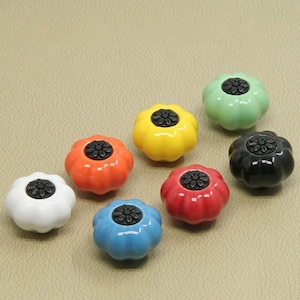 May include: Set of seven ceramic pumpkin-shaped drawer pulls in various colours, including white, yellow, orange, red, blue, green, and black. Each pull has a black knob in the centre.
