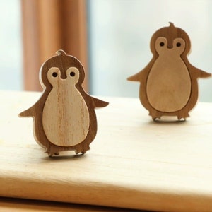 Peut inclure: Deux figurines de pingouin en bois, l'une face à la caméra et l'autre dos à la caméra. Les deux pingouins sont sculptés dans du bois brun clair et ont une finition en bois naturel.
