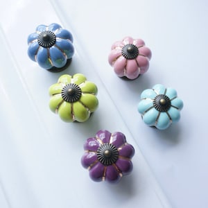 Op de afbeelding: Vijf keramische kastknoppen in verschillende kleuren, waaronder paars, groen, blauw, roze en lichtblauw. Elke knop heeft een gouden rand en een gouden midden met een reliëfdesign.