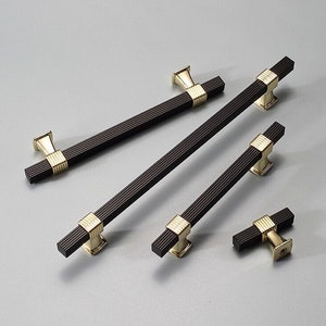 Pode incluir: Cinco puxadores de armário pretos e dourados com forma retangular e design estriado. Os puxadores são de vários tamanhos.