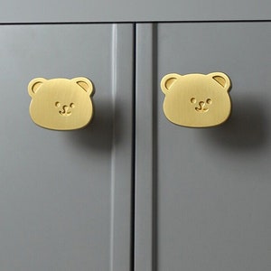 May include: Two gold bear-shaped cabinet knobs with a simple, smiling face design.