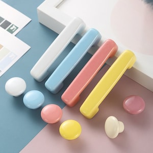 Puede incluir: Surtido de pomos y tiradores de cajones de cerámica de colores vivos en blanco, azul claro, rosa y amarillo. Los pomos son redondos y los tiradores son rectangulares.