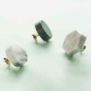 Puede incluir: Tres pomos de armario de mármol en diferentes formas: un pomo redondo, un pomo hexagonal y un pomo ovalado. Los pomos son blancos y grises con detalles dorados.