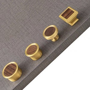 Könnte beinhalten: Vier Messing-Schrankknöpfe mit Holzeinlagen. Die Knöpfe sind rund und haben einen erhöhten Rand. Die Holzeinlagen sind dunkelbraun und haben eine natürliche Maserung.