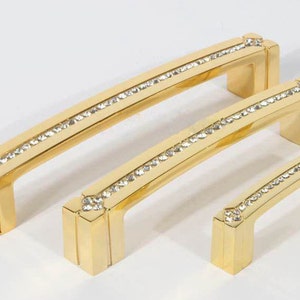 May include: Three gold cabinet pulls with a row of clear crystals around the edge. The pulls are different sizes.