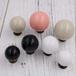 Könnte beinhalten: Sortiment von acht Keramik-Schrankknöpfen in verschiedenen Farben, darunter Schwarz, Weiß, Rosa und Beige. Die Knöpfe sind rund und haben eine glatte, glänzende Oberfläche.