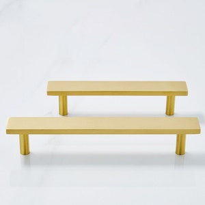 May include: Two gold-toned cabinet pulls with a rectangular shape and a brushed finish. The pulls are approximately 5 inches long.