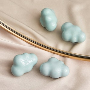 Op de afbeelding: Vier lichtblauwe, wolkvormige sculpturen van keramiek op een beige stoffen achtergrond.