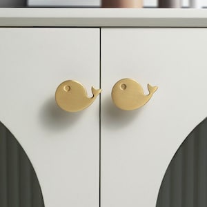 Könnte beinhalten: Zwei goldene, walförmige Schrankknöpfe sind an einer weißen Schranktür montiert. Die Schranktür hat eine abgerundete Kante.