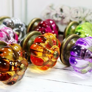 Peut inclure: Assortiment de boutons de cabinet en verre coloré en forme de citrouille avec une base en laiton. Les boutons sont de différentes couleurs, notamment le violet, le vert, l'orange, le rose et le marron.