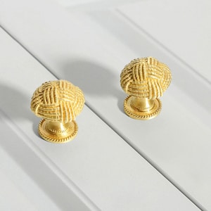 Peut inclure: Deux boutons de cabinet dorés avec un motif tissé. Les boutons sont ronds et ont un bord surélevé.