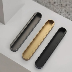 Könnte beinhalten: Drei Schrank-Schubladengriffe in verschiedenen Ausführungen: Schwarz, Gold und Grau. Jeder Griff hat eine längliche, rechteckige Form mit einem eingelassenen Griff.