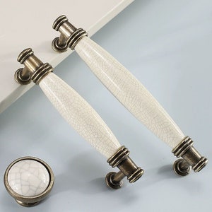 Puede incluir: Tres pomos de armario de bronce antiguo con manijas de cerámica blanca. Las manijas tienen un acabado craquelado. Los pomos son redondos y tienen un diseño decorativo.